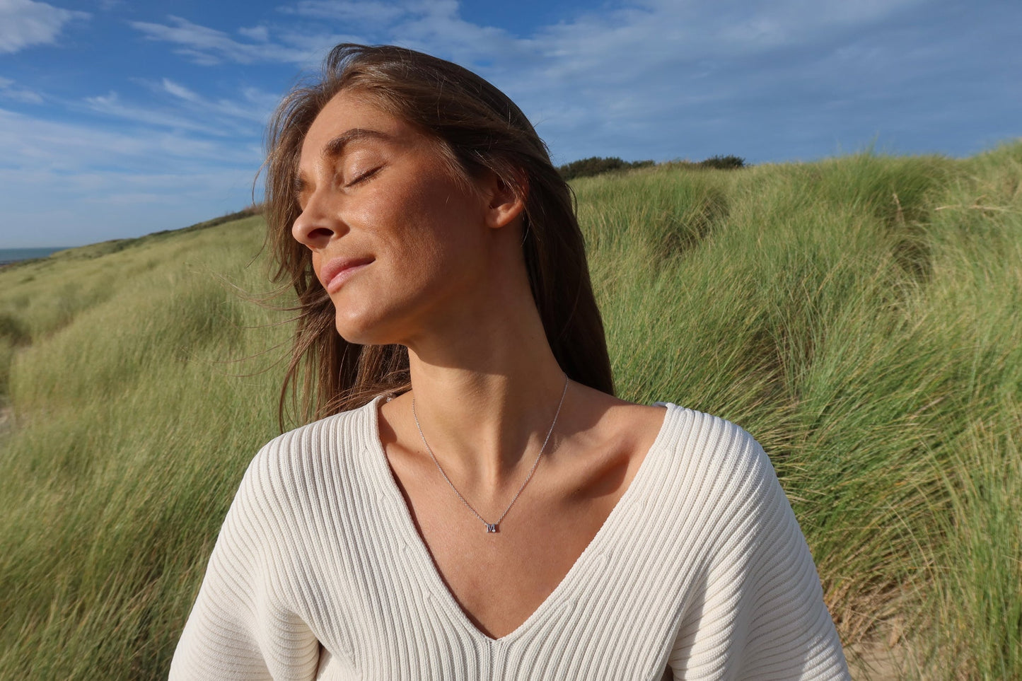 Une femme se tient dans un grand champ dherbe verte, les yeux fermés, portant un haut blanc et le collier « Anna | Initiale Lait Maternel Or » de Mama Jewels, son visage incliné vers le ciel partiellement nuageux, se prélassant au soleil.
