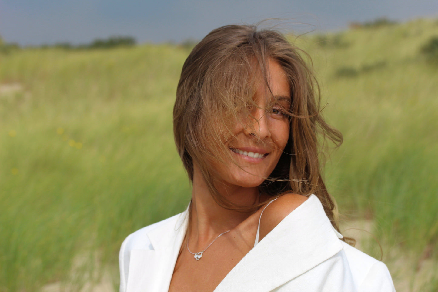 Une femme aux longs cheveux bruns sourit dans un champ herbeux, vêtue dun haut blanc tandis que ses cheveux flottent doucement sur son visage. Ce moment serein sapparente au lien unique symbolisé par le collier Lait Maternel Argent « Trinity » de Mama Jewels.