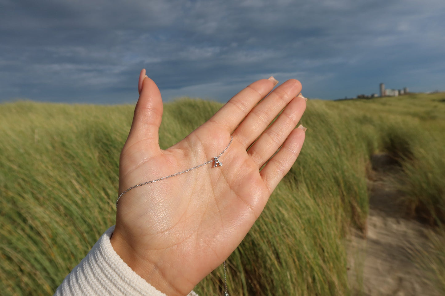 Une main aux ongles roses tient délicatement le collier « Anna | Initiale Lait Maternel Or » de Mama Jewels, son pendentif « R » en argent reflétant un charme unique sur fond dherbe haute et de ciel nuageux. Un chemin sablonneux menant à un bâtiment lointain capture lessence des bijoux uniques dans cette scène personnalisée.