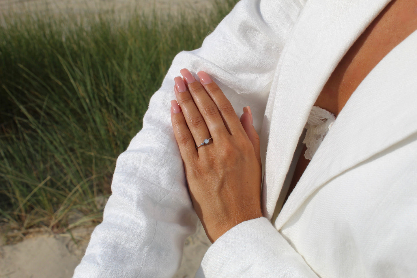 Une personne en tenue blanche met en valeur sa main avec une manucure rose et la Giulia | Bague Lait Maternel Argent de Mama Jewels, sur fond dherbe extérieure.