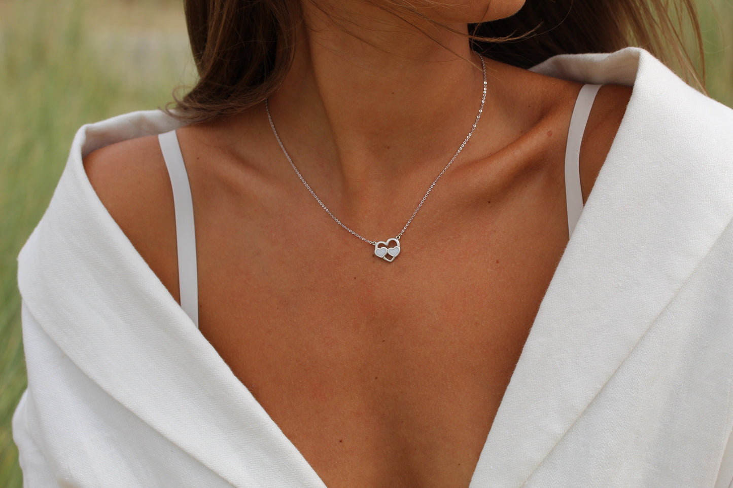 Une personne porte un haut blanc à épaules dénudées et le collier Trinity Lait Maternel Argent de Mama Jewels avec un pendentif en forme de cœur. Le collier est au centre de lattention, symbolisant votre lien unique avec votre enfant, tandis que le visage de la personne nest pas visible.
