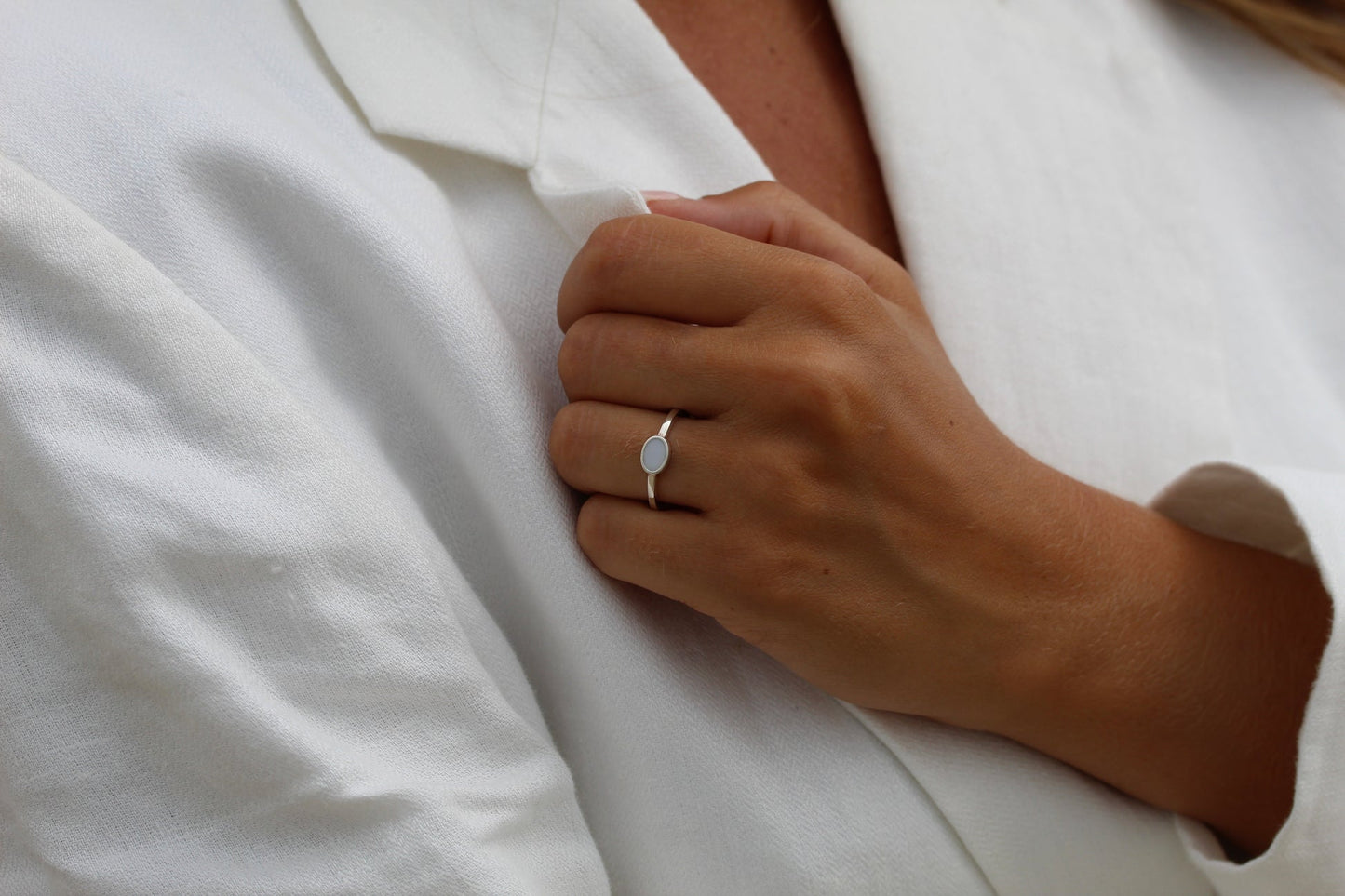 Une personne en blouse blanche tient le revers de sa veste avec sa main droite, dévoilant une bague Juna de Mama Jewels. La Bague Lait Maternel Or est dotée dune simple bande argentée et dune pierre blanche ovale qui capte la lumière avec son design captivant.