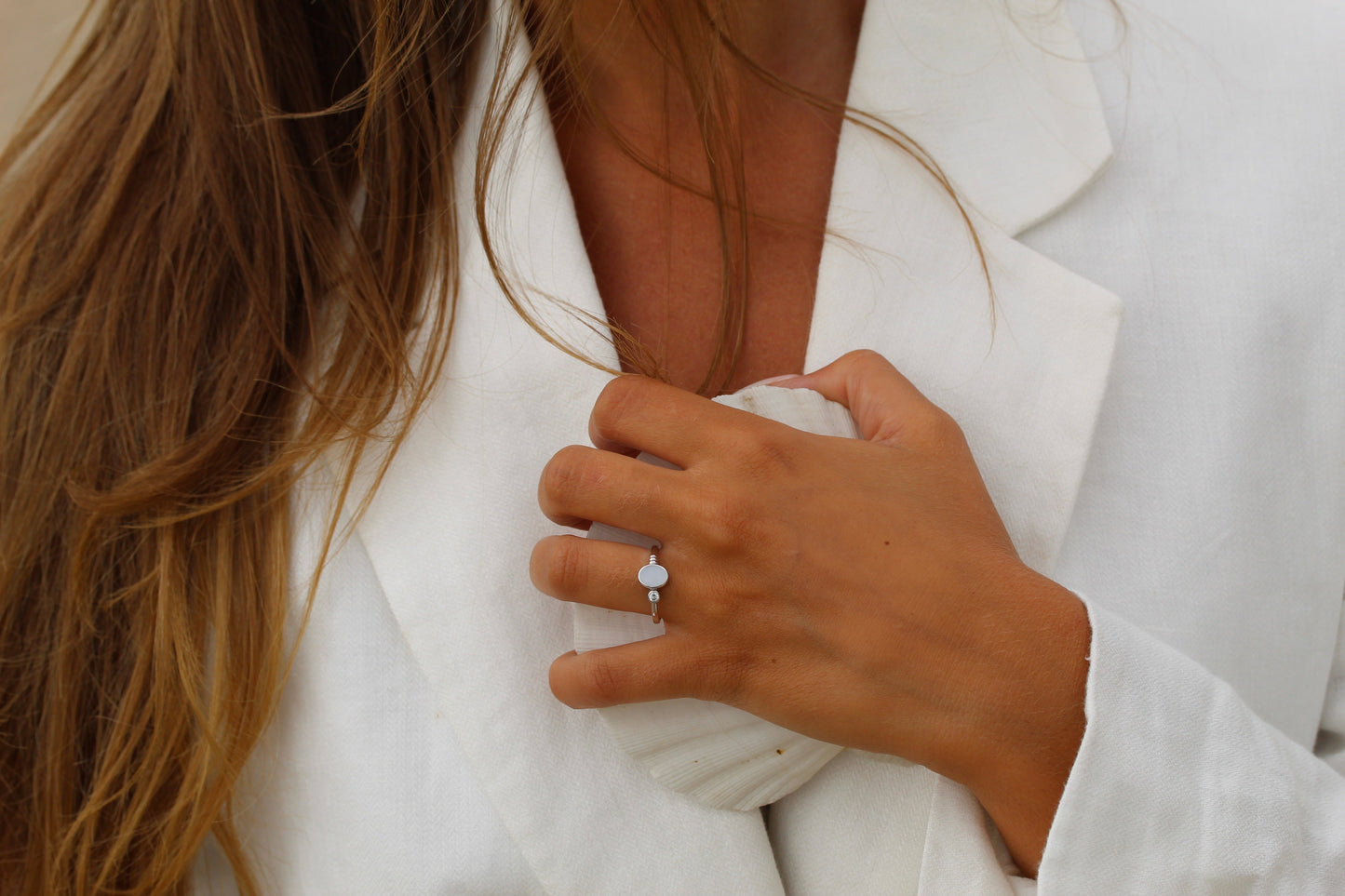 Une personne aux cheveux longs portant une veste blanche tient un coquillage contre sa poitrine, mettant en valeur lElisa de Mama Jewels, une saisissante Bague Lait Maternel Argent comportant une pierre circulaire blanche sur son doigt.