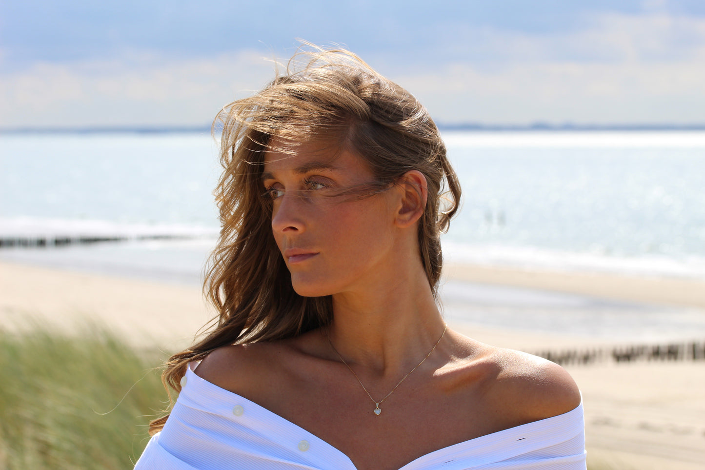 Une femme aux longs cheveux bruns se tient sur une plage, vêtue d'un haut blanc décolleté et du collier Cara | Pendentif Lait Maternel Argent de Mama Jewels. Ses cheveux balayés par le vent encadrent son visage alors qu'elle regarde sur le côté, avec la mer et le sable derrière elle.