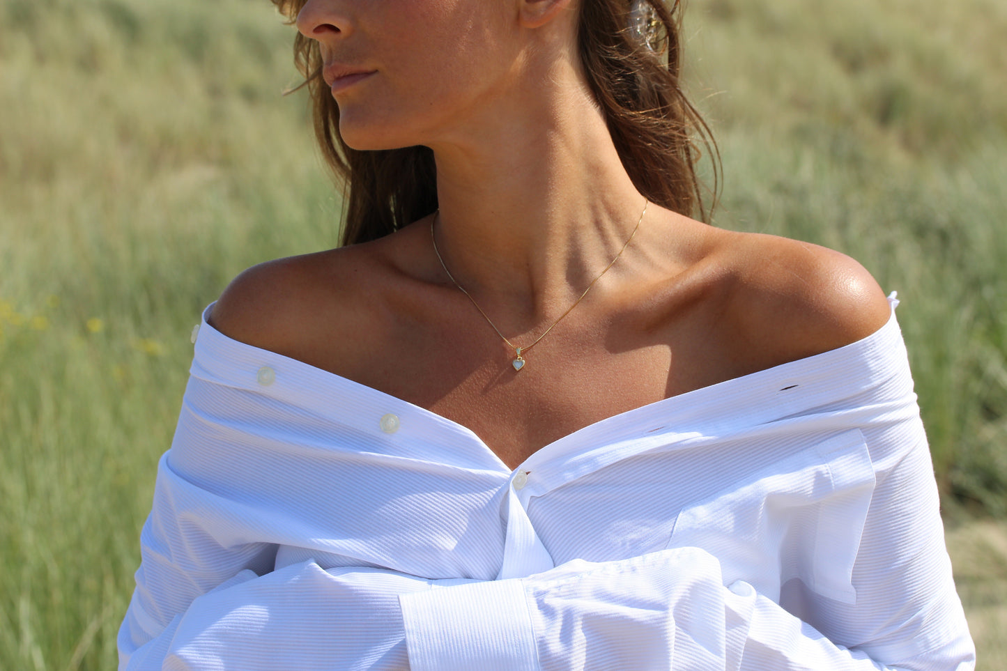 Une femme se tient dans un champ herbeux, ses cheveux flottant dans la brise. Elle porte une chemise blanche décolletée et le Cara | Pendentif Lait Maternel Argent de Mama Jewels. Son visage est partiellement hors champ.