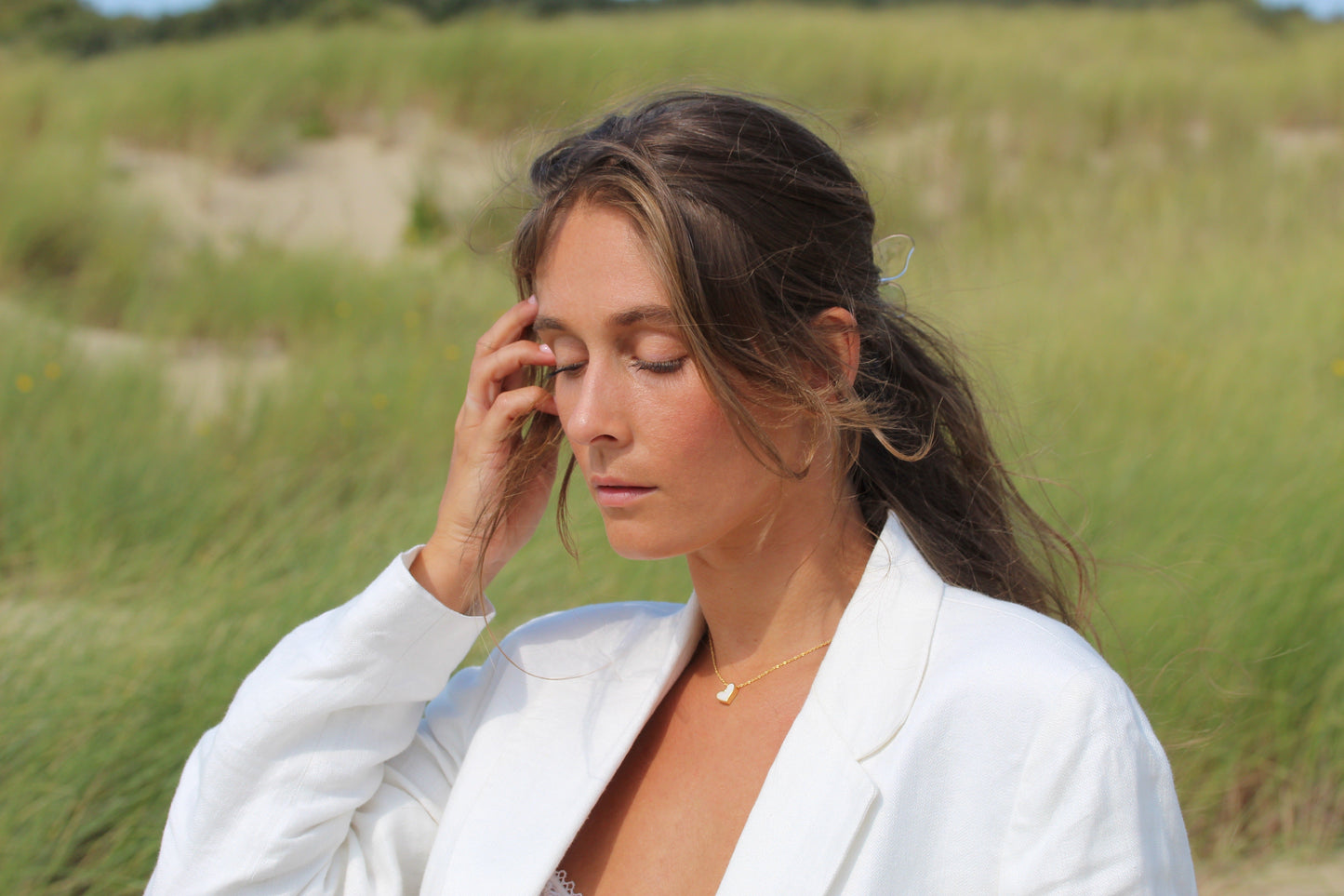 Dans un champ herbeux, une femme aux longs cheveux bruns attachés en queue de cheval se tient debout, les yeux fermés. Elle porte un blazer blanc et le collier Louisa Lait Maternel Argent de Mama Jewels, qui lui touche doucement le visage. Larrière-plan présente des dunes de sable et de la verdure sous un ciel clair.