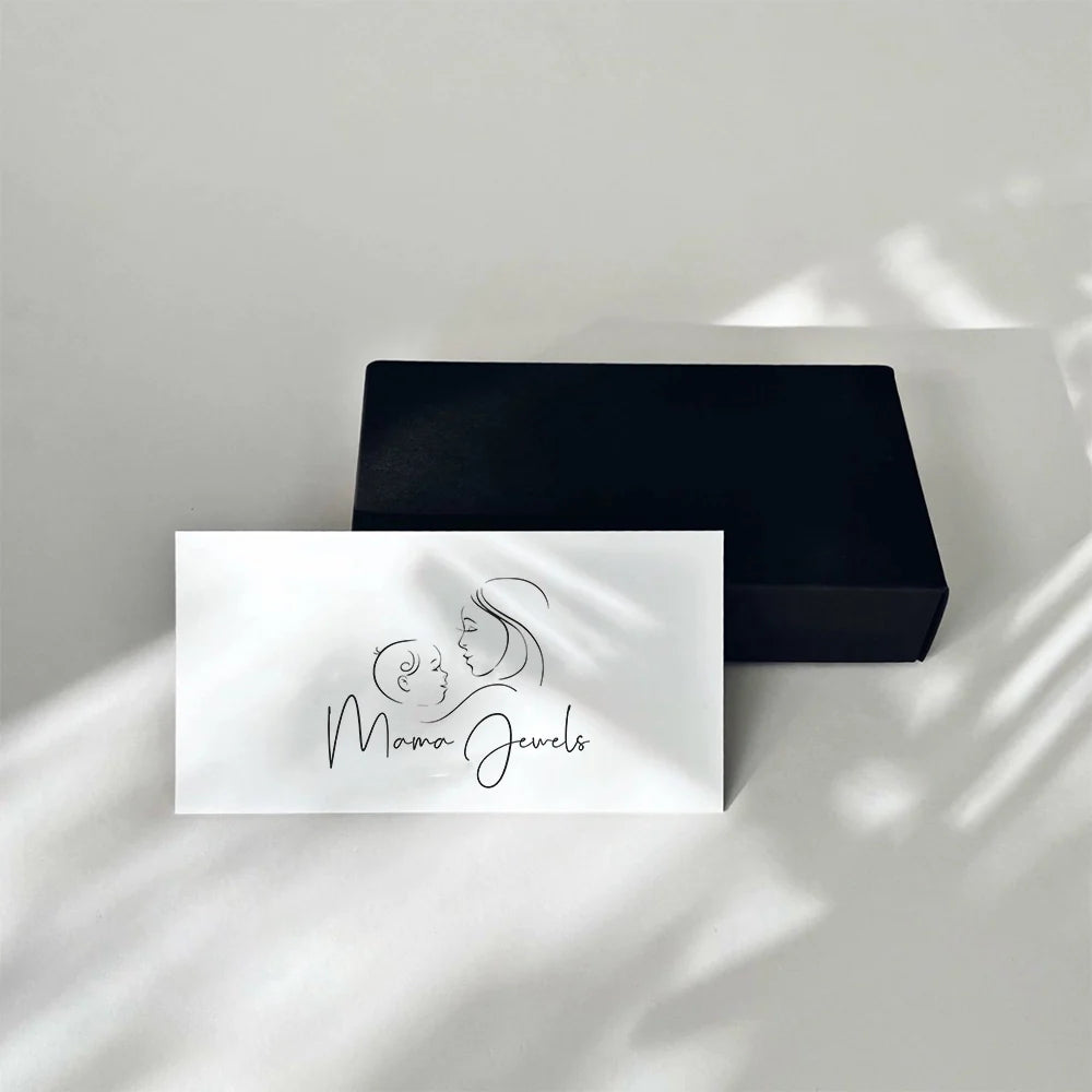 Une carte blanche ornée d'un dessin minimaliste représentant une mère et son enfant, ainsi que de l'inscription « Mama Jewels », repose sur une boîte noire fermée et unie. Le Chèque Cadeau Mama Jewels est généralement expédié sous 2 à 3 jours ouvrables.