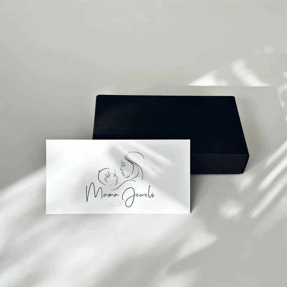 Une carte blanche représentant un dessin au trait minimaliste d'une femme et d'un enfant avec le texte « Mama Jewels » est exposée sur une boîte noire fermée — parfait pour accompagner le Chèque Cadeau Mama Jewels livré élégamment.