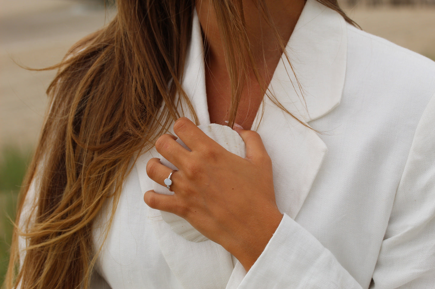 Une femme aux longs cheveux bruns portant un blazer blanc tient un coquillage contre sa poitrine, son doigt orné de la création unique de Mama Jewels : la bague Elisa | Bague Lait Maternel Or, dans un décor extérieur.
