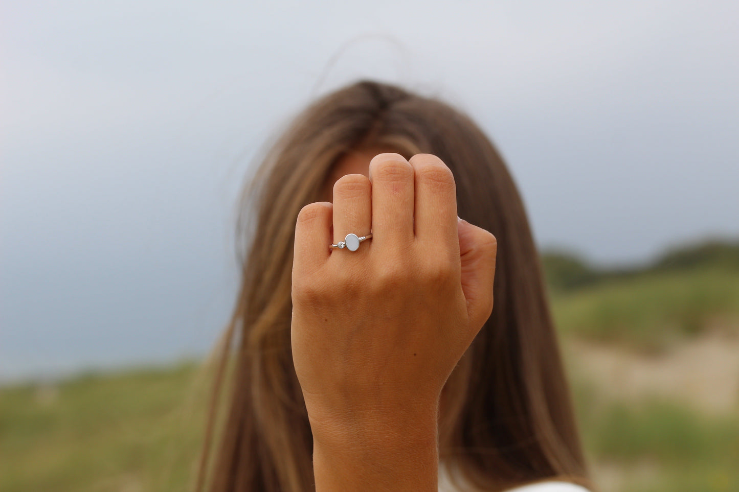 Une personne aux cheveux longs et bruns porte la bague en argent Elisa de Mama Jewels, ornée dune pierre claire. Le paysage flou, rempli de verdure et dun ciel nuageux, est en arrière-plan.