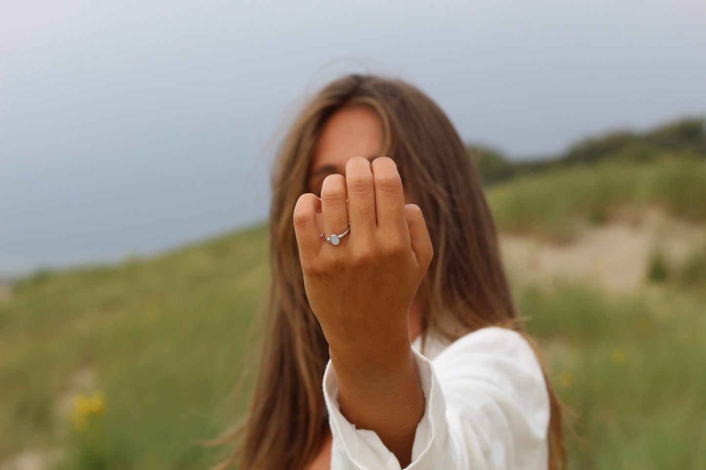 Une femme aux cheveux longs se tient à lextérieur contre des dunes herbeuses sous un ciel nuageux, mettant en valeur la bague Elisa | Bague Lait Maternel Or de Mama Jewels sur son doigt. Elle est vêtue dune chemise blanche.