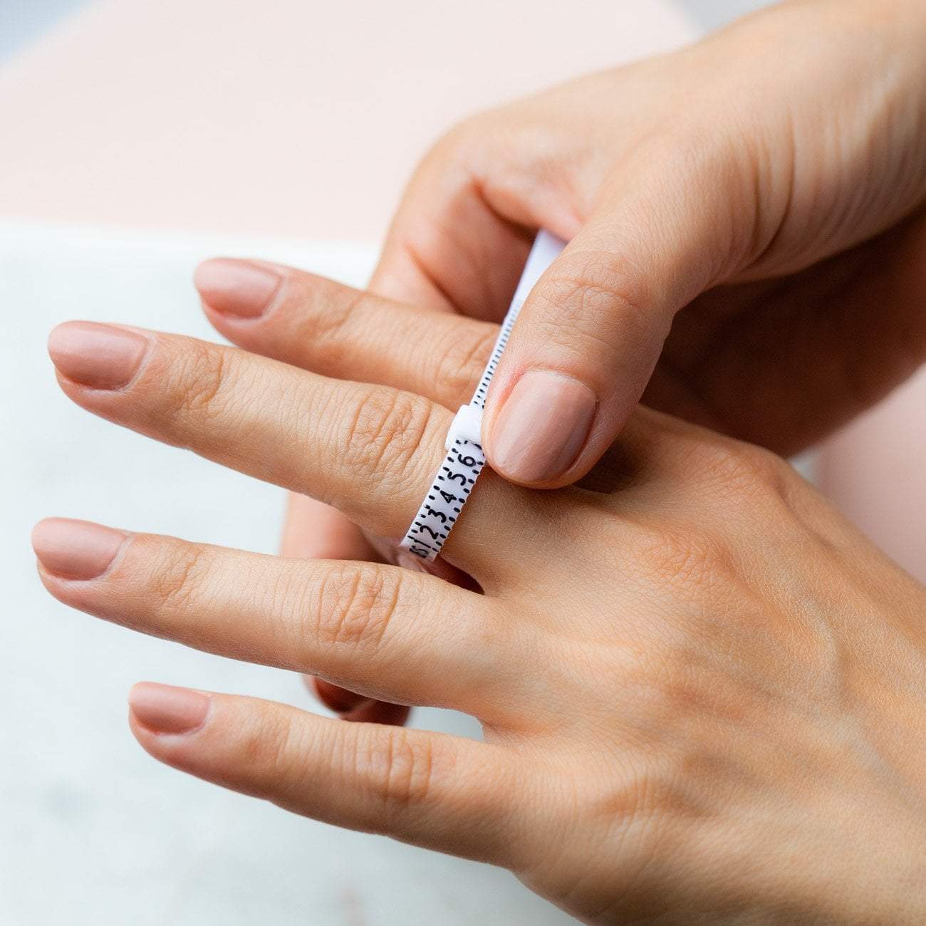 Une personne utilise la Bague d'ajustement pour une taille de bague parfaite de Mama Jewels avec un ruban blanc pour mesurer la circonférence du doigt, trouver sa taille de bague exacte et obtenir une réduction de 5 €.