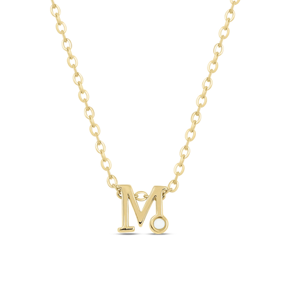 Le collier Anna | Initiale Lait Maternel Or de Mama Jewels est un collier chaîne en or avec un pendentif « M » personnalisé, orné dune petite pierre blanche ronde en bas à droite. Le fond noir uni met en valeur cette pièce unique.