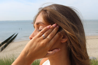 Une personne aux longs cheveux bruns et portant une bague Sofia de Mama Jewels au doigt tient la tête, les yeux fermés, face à la mer. La plage et locéan sétendent sous un ciel clair, offrant tranquillité et réflexion.