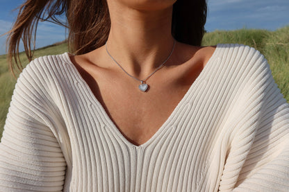 Une femme porte un pull côtelé blanc et le pendentif Mama Jewels Demi, un collier en argent en forme de cœur. Larrière-plan montre de lherbe verte et un ciel bleu, indiquant un décor extérieur.