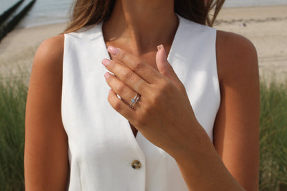 Sur une plage, une femme vêtue dun haut blanc sans manches met en valeur ses ongles vernis et la Bague Lait Maternel Argent de Mama Jewels Sofia sur fond de sable, dherbe et de mer.