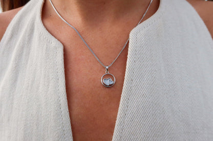 Un gros plan montre une personne portant le pendentif Amélie | Lait Maternel Argent de Mama Jewels. Le pendentif présente un logo Apple à lintérieur dun cadre circulaire, complétant son haut texturé avec un décolleté profond et dégageant une élégance.