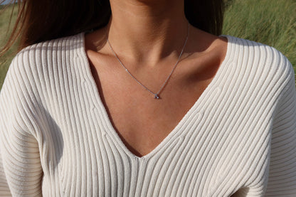 Une personne porte un pull blanc côtelé et le délicat collier en argent « Anna | Initiale Lait Maternel Or » de Mama Jewels, avec un petit pendentif personnalisé. Larrière-plan présente des éléments verts flous.
