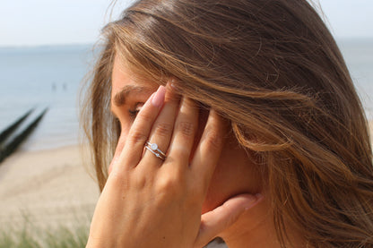 Une femme aux longs cheveux bruns touche son visage, mettant en valeur la bague Sofia | Bague Lait Maternel Argent de Mama Jewels sur son doigt. Elle se tient près dune plage, avec locéan et le sable visibles en arrière-plan.