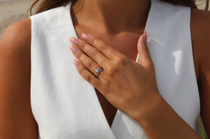 Une personne aux cheveux longs, vêtue dun haut blanc sans manches, pose délicatement une main sur sa poitrine. Elle a les ongles manucurés et porte lélégante Sofia | Bague Lait Maternel Argent de Mama Jewels ornée dun petit diamant sur lannulaire.