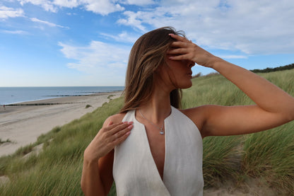 Une femme en haut blanc se tient debout sur une dune herbeuse avec la plage et locéan derrière elle. Elle se protège les yeux dune main tout en touchant son collier Mama Jewels Amélie | Pendentif Lait Maternel Argent de lautre. Le ciel est partiellement nuageux.