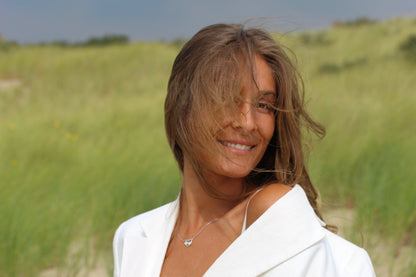 Une femme aux longs cheveux bruns sourit dans un champ herbeux, vêtue dun haut blanc tandis que ses cheveux flottent doucement sur son visage. Ce moment serein sapparente au lien unique symbolisé par le collier Lait Maternel Argent « Trinity » de Mama Jewels.