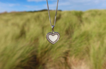 Le Demi | Pendentif Lait Maternel Argent de Mama Jewels est un bijou en argent sterling avec un pendentif en forme de cœur, doté dun centre blanc et dune bordure sertie de pierres précieuses. Il est élégamment suspendu à une chaîne délicate sur fond dherbe verte floue et de ciel bleu.