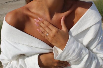 Une personne portant un haut blanc à épaules dénudées présente une bague Giulia | Bague Lait Maternel Argent de Mama Jewels et une bague en diamant, la main près de la poitrine. Ses ongles rose clair manucurés complètent le décor extérieur.