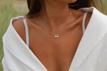 Une personne porte un haut blanc à épaules dénudées et le collier Trinity Lait Maternel Argent de Mama Jewels avec un pendentif en forme de cœur. Le collier est au centre de lattention, symbolisant votre lien unique avec votre enfant, tandis que le visage de la personne nest pas visible.