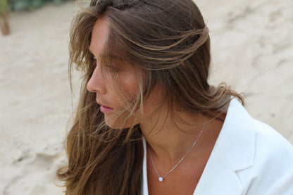 Sur une plage de sable, une femme aux cheveux bruns et vêtue dun haut blanc est assise sereinement, ses cheveux flottant doucement sur son visage. Elle porte le collier Lait Maternel Or Lis | de Mama Jewels, un délicat pendentif en forme de lait maternel qui capture un lien spécial, soulignant son expression pensive au milieu de la scène sereine.