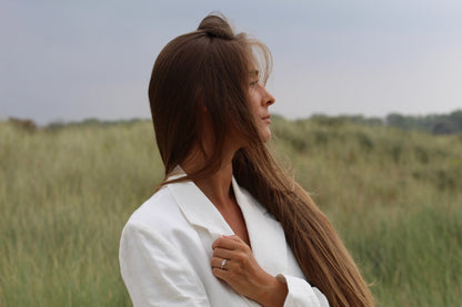 Dans un champ herbeux, une femme aux longs cheveux bruns porte un blazer blanc et la bague Juna, Bague Lait Maternel Argent de Mama Jewels. Elle regarde sereinement vers sa droite sous un ciel couvert avec une végétation luxuriante à lhorizon.