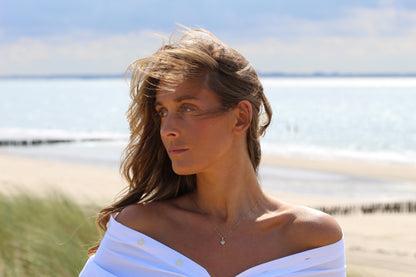 Une femme aux longs cheveux bruns se tient sur une plage, vêtue d'un haut blanc décolleté et du collier Cara | Pendentif Lait Maternel Argent de Mama Jewels. Ses cheveux balayés par le vent encadrent son visage alors qu'elle regarde sur le côté, avec la mer et le sable derrière elle.