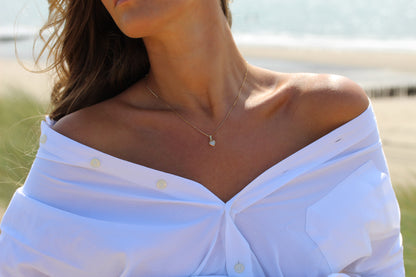 Une femme se tient à l'extérieur, près du sable et de l'eau. Elle porte une chemise blanche décolletée et le collier Mama Jewels Cara | Pendentif Lait Maternel Argent.