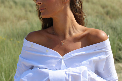 Une femme se tient dans un champ herbeux, ses cheveux flottant dans la brise. Elle porte une chemise blanche décolletée et le Cara | Pendentif Lait Maternel Argent de Mama Jewels. Son visage est partiellement hors champ.