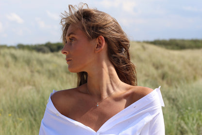 Une femme aux cheveux châtain clair se tient dans un champ herbeux, vêtue d'une chemise blanche décolletée et du collier Cara | Pendentif Lait Maternel Argent de Mama Jewels. L'herbe verte et un ciel partiellement nuageux sont à l'arrière-plan.