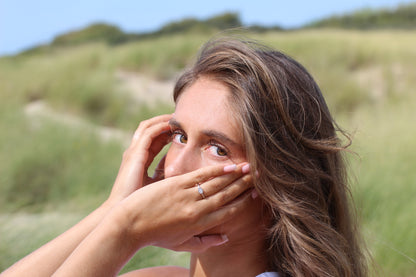 Une femme aux longs cheveux bruns se couvre la moitié du visage avec ses mains, affichant la superbe bague Luna | Bague Lait Maternel Argent de Mama Jewels. Elle se tient à lextérieur sur un fond vert et bleu flou, ressemblant à de lherbe et du ciel.