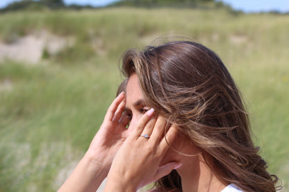 Une femme aux longs cheveux bruns se touche les tempes des deux mains alors quelle se tient debout à lextérieur, où lherbe verte floue et le ciel bleu lui servent de toile de fond. Sur sa main gauche, une Bague Lait Maternel Argent Luna | de Mama Jewels scintille au soleil.