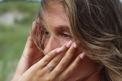 Une personne aux longs cheveux bruns se couvre légèrement les yeux avec ses mains aux ongles rose clair. Une main porte la bague Luna de Mama Jewels, en argent sterling. Larrière-plan présente une verdure floue.
