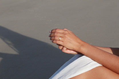 Gros plan de mains posées sur une surface claire avec la lumière du soleil projetant une ombre. La personne porte la bague Luna | Bague Lait Maternel Or de Mama Jewels, renforçant son charme sur le fond lisse et neutre.
