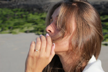 Une femme aux longs cheveux bruns pose à l'extérieur, présentant la Luna | Bague Lait Maternel Or de Mama Jewels, avec une pierre de lait maternel, avec du sable et de la verdure doucement flous en arrière-plan.