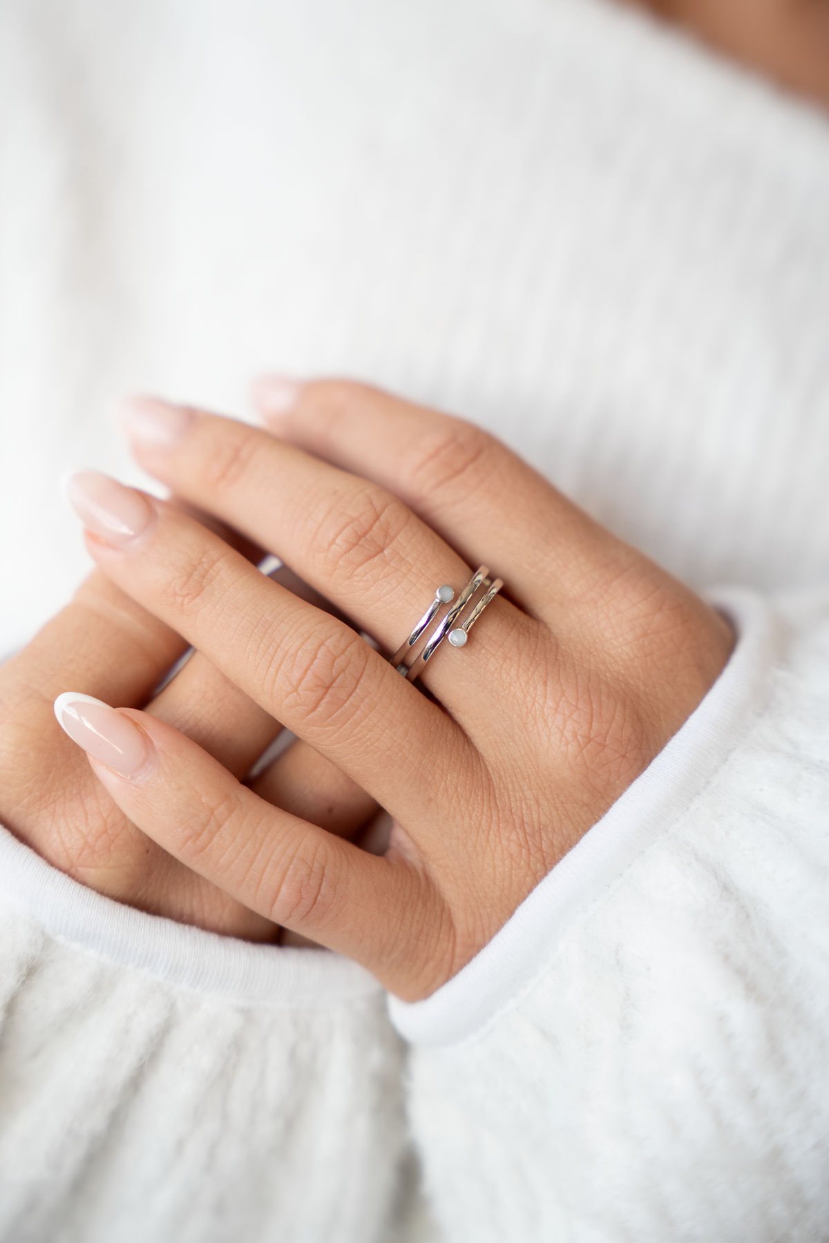 Bague Aurore en argent portée à l'annulaire, spirale ouverte et deux petites pierres de lait claires, pull blanc