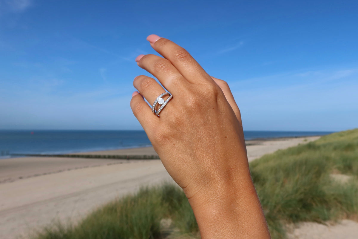 Main levée devant la plage portant une large bague argentée à plusieurs anneaux et pierre de lait ronde