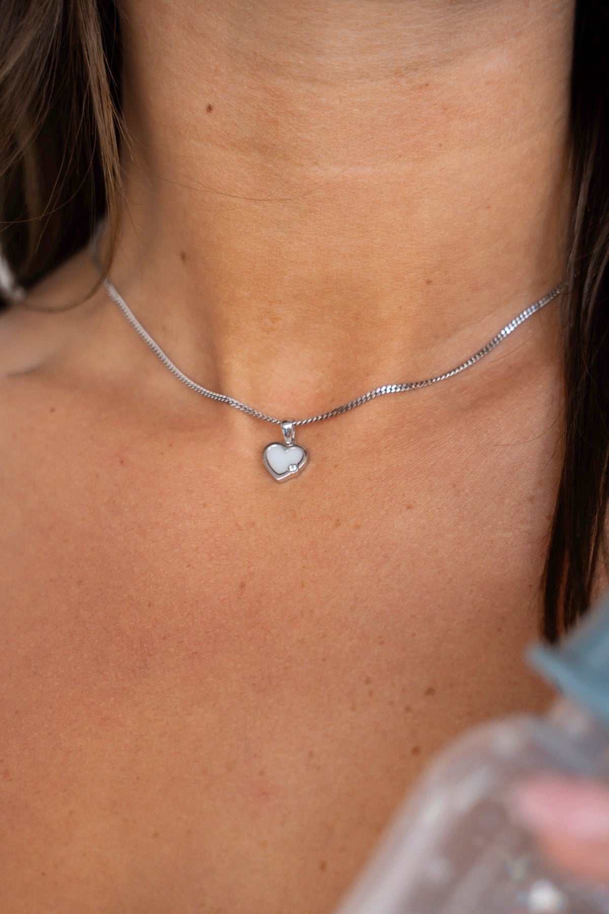 Gros plan sur le pendentif Esmée en or blanc avec cœur de lait maternel sur la clavicule, à côté d'un biberon de lait.