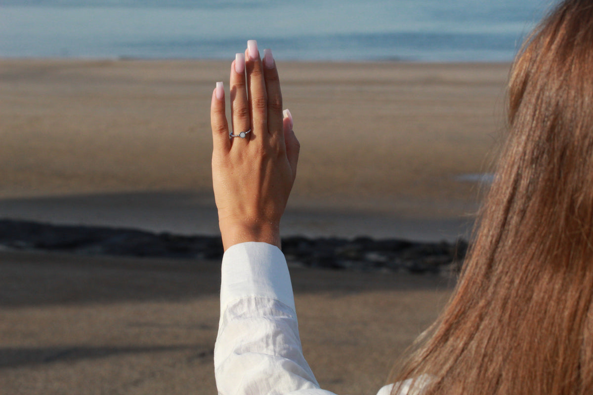 Main levée devant la mer portant la bague Isabelle en argent, manche de chemise blanche