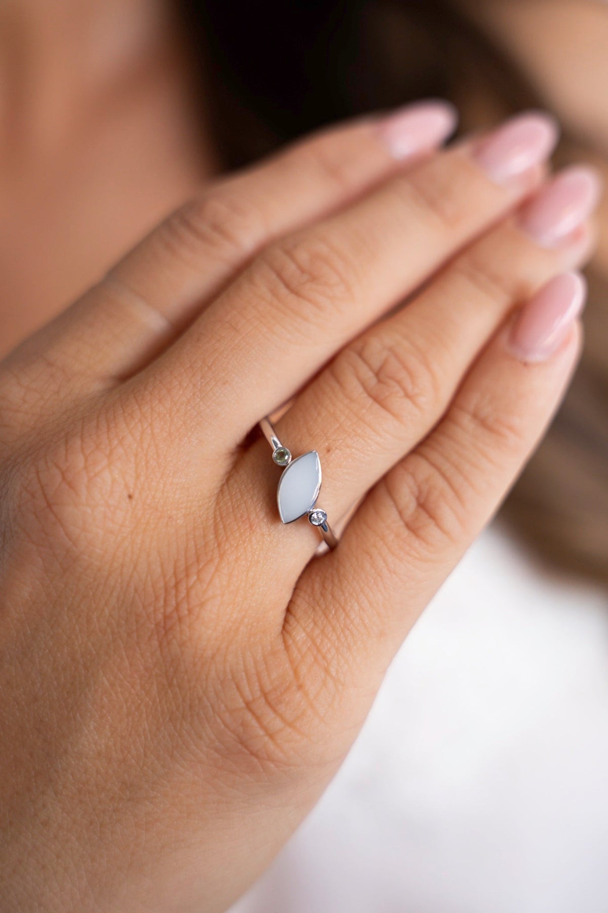 Gros plan de la bague Lina Duo en or blanc : une pierre ovale de lait maternel entre deux petites pierres de naissance, portée à la main.