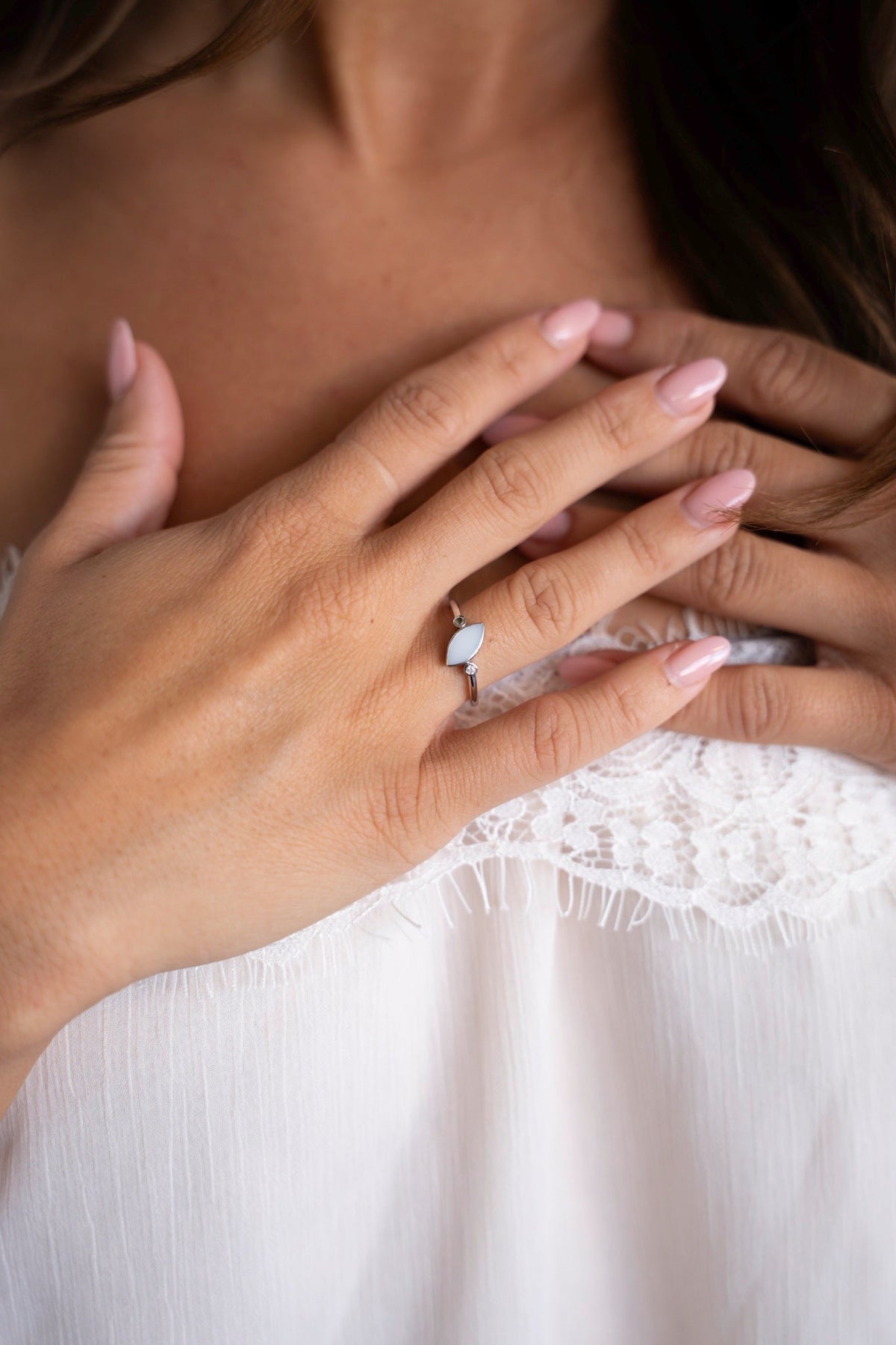 Mains sur la poitrine avec la bague Lina Duo en or blanc, une pierre de lait maternel et deux petites pierres, devant un haut en dentelle blanc.
