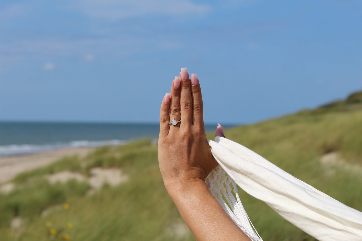 Main levée devant la mer et les dunes portant la bague Turning Droplet avec goutte de lait maternel