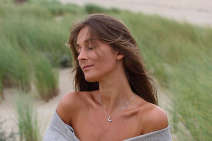 Une femme aux cheveux longs ferme les yeux, savourant la brise. Elle porte Ayla | Pendentif Lait Maternel Or de Mama Jewels, un pendentif unique en forme de croissant qui scintille à la lumière, et un châle léger drape ses épaules. Derrière elle se trouvent des dunes de sable et de hautes herbes.
