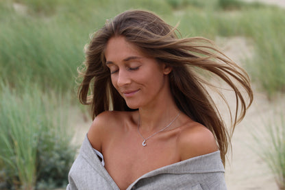 Une femme aux longs cheveux bruns se tient dehors, les yeux fermés et souriant doucement, portant un haut gris à épaules dénudées et le collier Ayla | Pendentif Lait Maternel Or de Mama Jewels. Le vent ébouriffe ses cheveux tandis que lherbe verte décore larrière-plan flou.