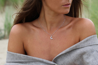 Une femme aux cheveux longs bruns porte le pendentif Lait Maternel Or Ayla de Mama Jewels, un collier unique en argent en forme de croissant de lune qui capte la lumière. Elle est enveloppée dans un tissu texturé gris, ses épaules exposées sur un fond flou de verdure et de sable.